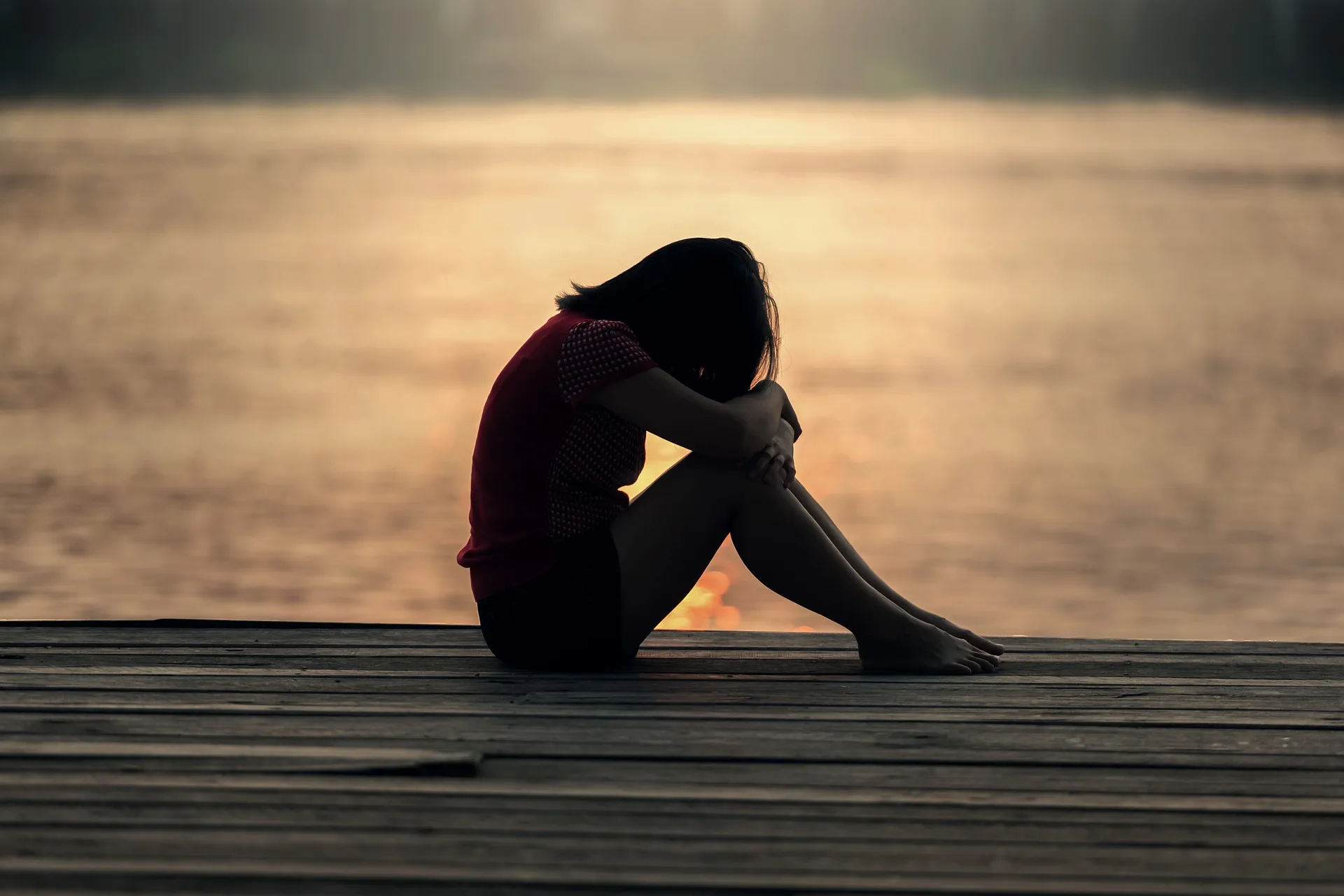 A woman sitting grieving with her head on her knees on a boardwalk at sunset.