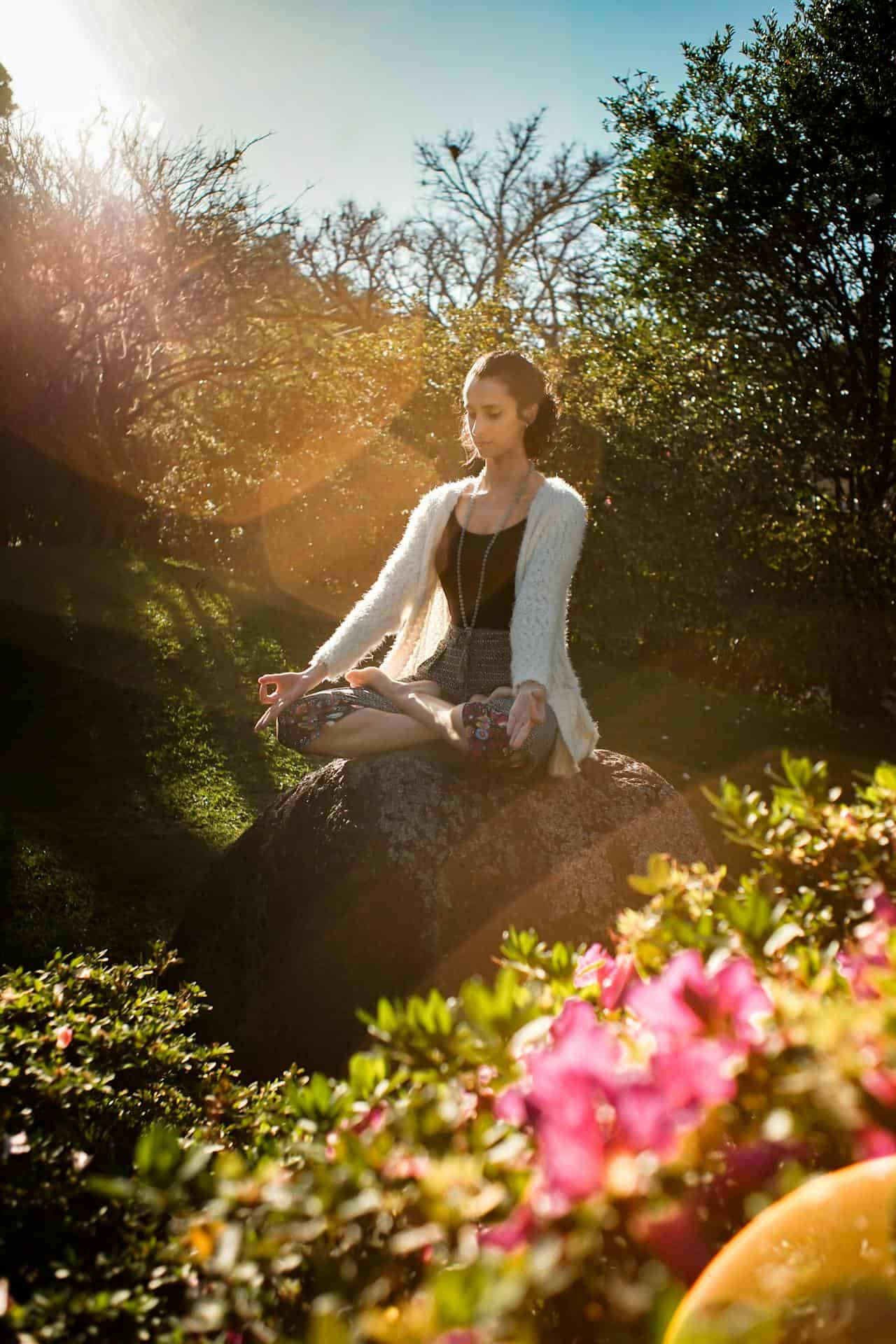 A woman meditating in peace on top of a rock surrounded by floral nature.